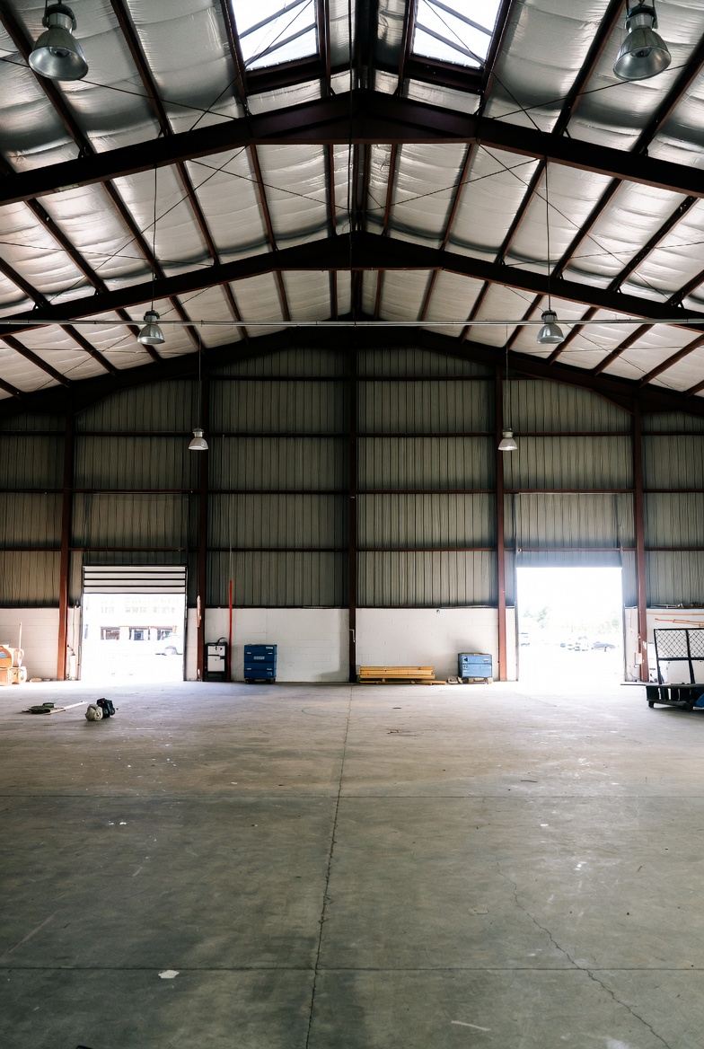 Indvendigt foto af en stor, tom lagerhal med beton­gulv, højt metal­tag og åbne porte, der giver naturligt lys ind i rummet.